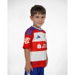 Primera equipación oficial del Granada CF para niños, temporada 2026-27. Conjunto de camiseta y pantalón rojiblancos.